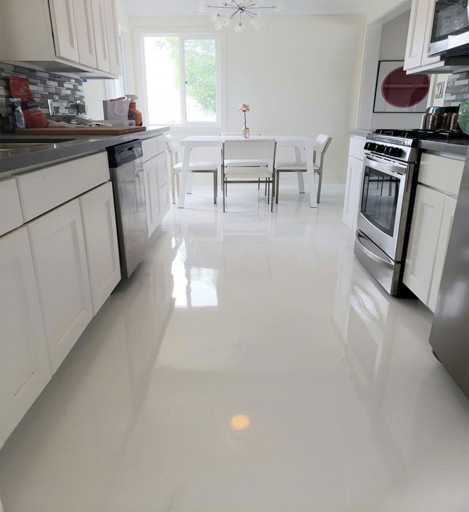 A white kitchen, with cuntertops in the left and right side, a table and chairs at the end, and a shiny white epoxy floor.